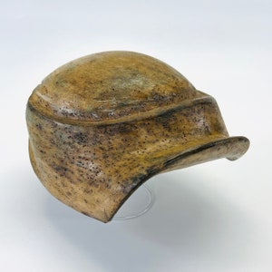 Könnte beinhalten: Ein brauner Keramikhelm mit einer strukturierten Oberfläche und einem gebogenen Rand. Der Helm hat ein abgenutztes, gealtertes Aussehen.