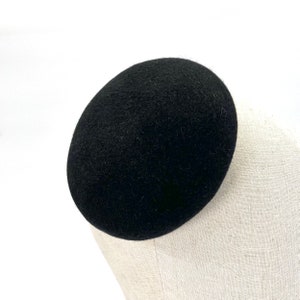 May include: A black felt pillbox hat. The hat is round and has a smooth, slightly textured surface.