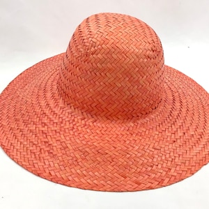 Könnte beinhalten: Ein breitkrempiger Strohhut in hellkorallenfarbener Farbe. Der Hut ist mit einem einfachen Muster gewebt und hat eine leicht abgerundete Krone.