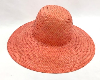 Puede incluir: Un sombrero de paja de ala ancha de color coral claro. El sombrero está tejido con un patrón simple y tiene una corona ligeramente redondeada.