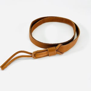 Peut inclure: Une lanière en cuir marron clair enroulée, avec un détail noué et deux extrémités pendantes. La lanière est faite d'une seule pièce de cuir et semble souple. Le fond est blanc uni.