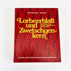 Può includere: Un libro con copertina rigida con una copertina a motivo a maglia rossa. Il titolo "Lorbeerblatt und Zwetschgenkern" è in grandi lettere gialle. Il libro include anche testo in tedesco, compresi i nomi degli autori e il nome dell'editore.