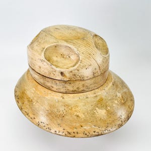 Peut inclure: Un bloc à chapeau en bois beige clair avec une calotte arrondie lisse présentant trois indentations circulaires sur la surface supérieure et un large bord évasé. Le bois affiche un grain naturel avec des taches sombres et une finition polie.