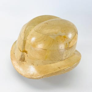 Könnte beinhalten: Eine Vintage-Hutform aus hellem Holz. Die Hutform besteht aus mehreren gebogenen Holzstücken, die eine abgerundete Krone und einen Rand bilden. Das Holz hat ein natürliches, gealtertes Aussehen.