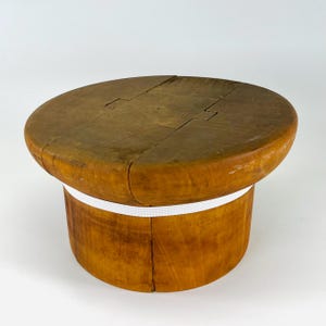 Könnte beinhalten: Ein Vintage-Hutblock aus Holz mit rundem Oberteil und zylindrischem Sockel. Der Hutblock ist aus hellbraunem Holz gefertigt und hat ein weißes Gummiband um den Sockel. Die Oberseite besteht aus mehreren Holzstücken, die miteinander verbunden sind.