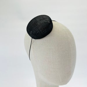 May include: A small, round, black fascinator hat with a textured, woven surface. The hat is secured to a thin, black headband that curves around the head form, positioning the circular piece at a slight angle on the top side of the head.