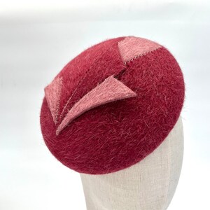 Puede incluir: Un sombrero de fieltro burdeos con un acento de fieltro rosa. El sombrero tiene una textura peluda y una forma redondeada.