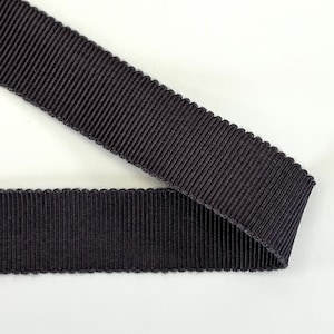 Pode incluir: Close-up de um pedaço de fita de gorgorão preta. A fita tem uma textura canelada com uma borda ondulada. A fita está dobrada sobre si mesma, mostrando ambos os lados do material. O fundo é branco.