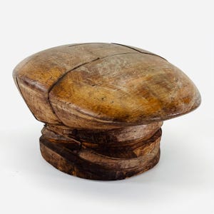 May include: A vintage wooden hat block with a rounded top and a layered base. The hat block is made of light and dark brown wood, showing visible grain and wear. The hat block is likely used for shaping hats.