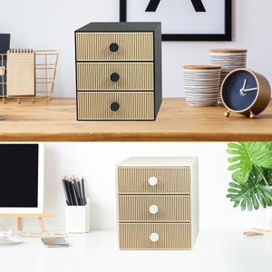 May include: A three-drawer storage unit with a black frame and light wood-toned drawer fronts. Each drawer has a black knob. The drawers have a vertical, ribbed design. The unit is on a wooden desk.