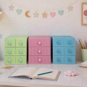 May include: Three small, colorful plastic drawer units in pastel shades of green, pink, and blue. Each unit has six drawers with round knobs. A notebook and pencil are in the foreground, with decorative wall hangings in the background.