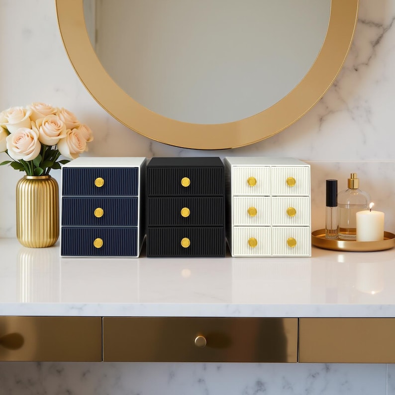 May include: Three small, decorative drawer sets in navy blue, black, and white, each with three drawers and gold-colored knobs. The sets are arranged on a white surface, with a gold-framed mirror in the background. A vase of flowers and a tray with perfume and a candle are also visible.