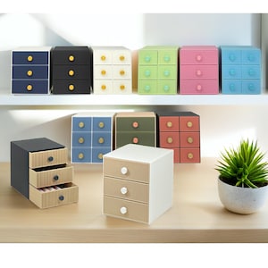 Puede incluir: Una colección de pequeños cajones de almacenamiento coloridos con tres o seis compartimentos. Los cajones son de varios colores, incluyendo azul, negro, blanco, verde, rosa y beige. Cada cajón tiene un pomo de color contrastante. También se ve una pequeña planta en maceta.