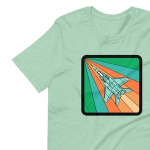 Puede incluir: Una camiseta gráfica de estilo retro con un avión de reacción azul volando a través de un arcoíris de colores. El diseño está sobre un fondo verde claro.
