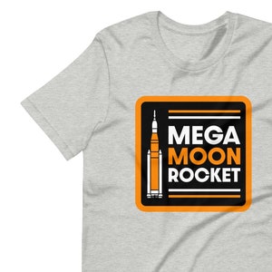 May include: A light gray t-shirt featuring a graphic with the words "MEGA MOON ROCKET" in white and orange, with a rocket ship illustration. The graphic is in a black and orange square.
