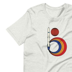 Puede incluir: Una camiseta blanca con un gráfico de un cohete espacial volando a través de un anillo de planetas coloridos. El cohete es blanco con detalles azules y los planetas son rojos, naranjas, amarillos y azules.