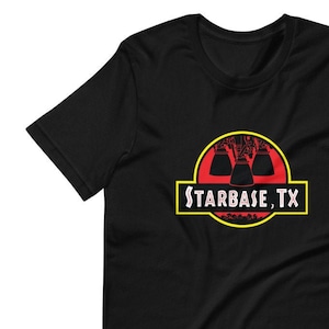 Könnte beinhalten: Schwarzes T-Shirt mit einer roten und gelben Grafik eines Dinosauriers mit drei Raketenmotoren im Inneren. Der Text "STARBASE, TX" befindet sich unter dem Dinosaurier.