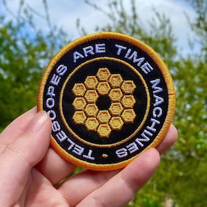 Può includere: Una toppa ricamata nera e dorata con il testo "I telescopi sono macchine del tempo" in un cerchio. La toppa presenta un design a nido d'ape al centro.