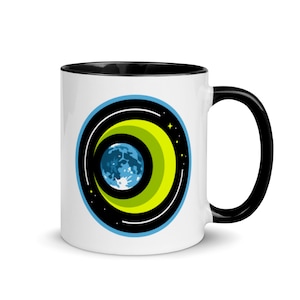 May include: A white ceramic coffee mug with a black rim and handle. The mug features a graphic of a blue and white moon surrounded by a black and yellow ring with a star.