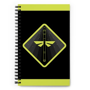 May include: A black and yellow spiral notebook with a dragonfly design on the cover. The dragonfly is inside a yellow diamond shape with a black background. The notebook has a black spiral binding.