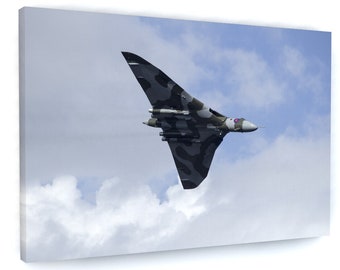 May include: A gray and green camouflage military aircraft flying in a blue sky with white clouds. The aircraft has a long, narrow fuselage and swept wings.