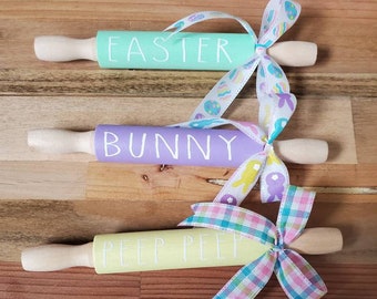 May include: Four wooden rolling pins decorated with pastel colors and ribbons. The rolling pins are labeled "Easter", "Bunny", "Peep Peep", and "Happy Easter".
