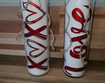May include: Two glass candle holders filled with white wax. One has "xoxo" in red glitter, the other has "love" in red glitter. Both are wrapped with twine and topped with a red heart gem.