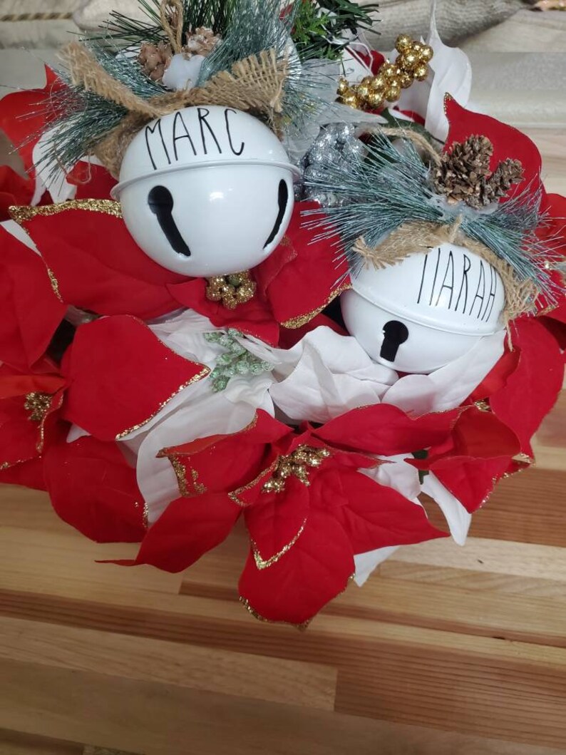 May include: A Christmas wreath with red poinsettia flowers and two white bells with the names "Marc" and "Marah" written on them.
