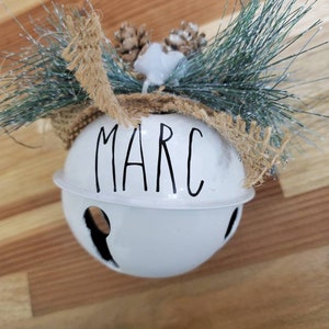 May include: White metal jingle bell ornament with the name "MARC" written in black lettering. The bell is decorated with a brown burlap bow and a sprig of faux pine.