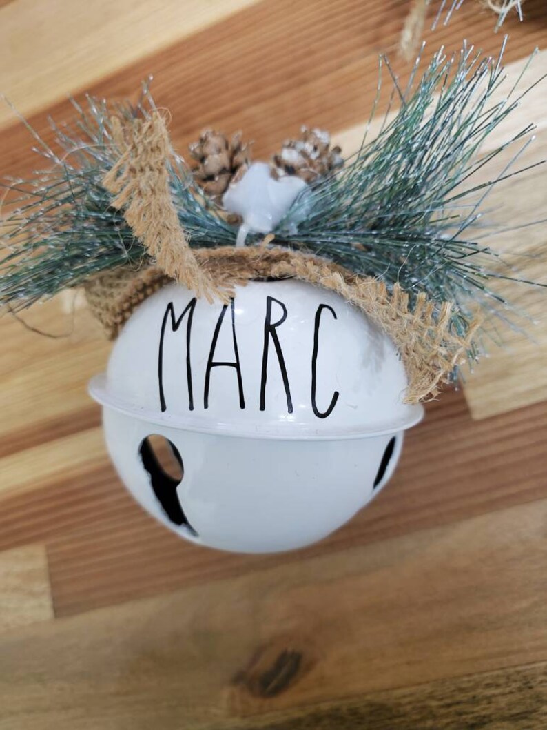 May include: White metal jingle bell ornament with the name "MARC" written in black lettering. The bell is decorated with a brown burlap bow and a sprig of faux pine.