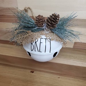 May include: White metal bell ornament with the name "Brett" written in black. The bell is decorated with a burlap bow and two brown pine cones.