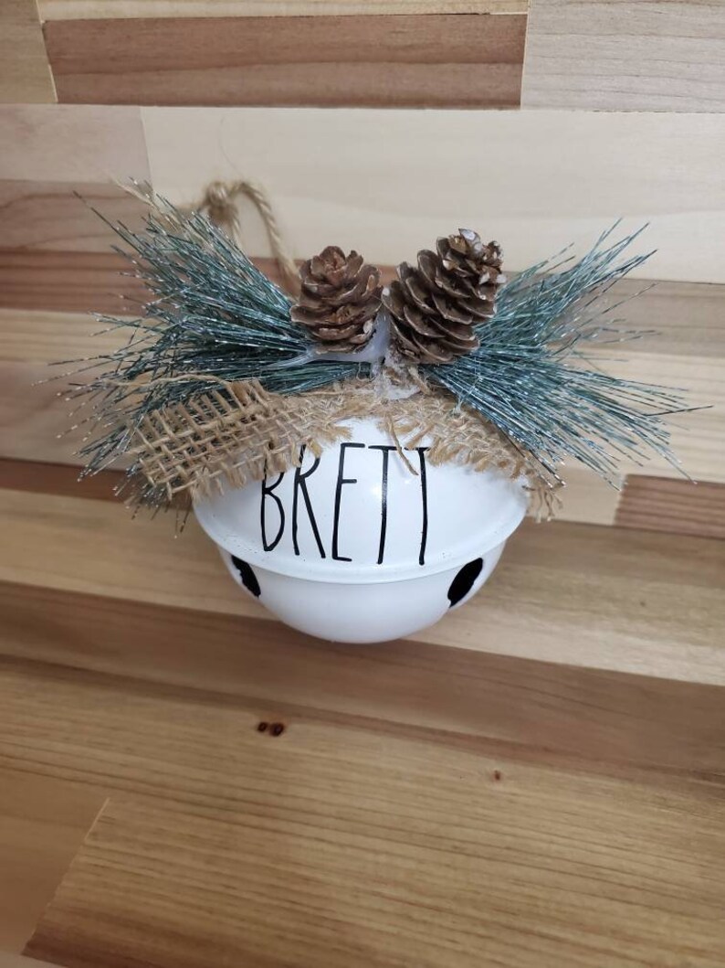 May include: White metal bell ornament with the name "Brett" written in black. The bell is decorated with a burlap bow and two brown pine cones.