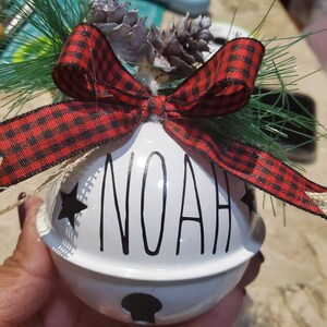 May include: White ceramic jingle bell ornament with the name "NOAH" written in black script. The bell is decorated with a red and black plaid bow and two small pine cones.