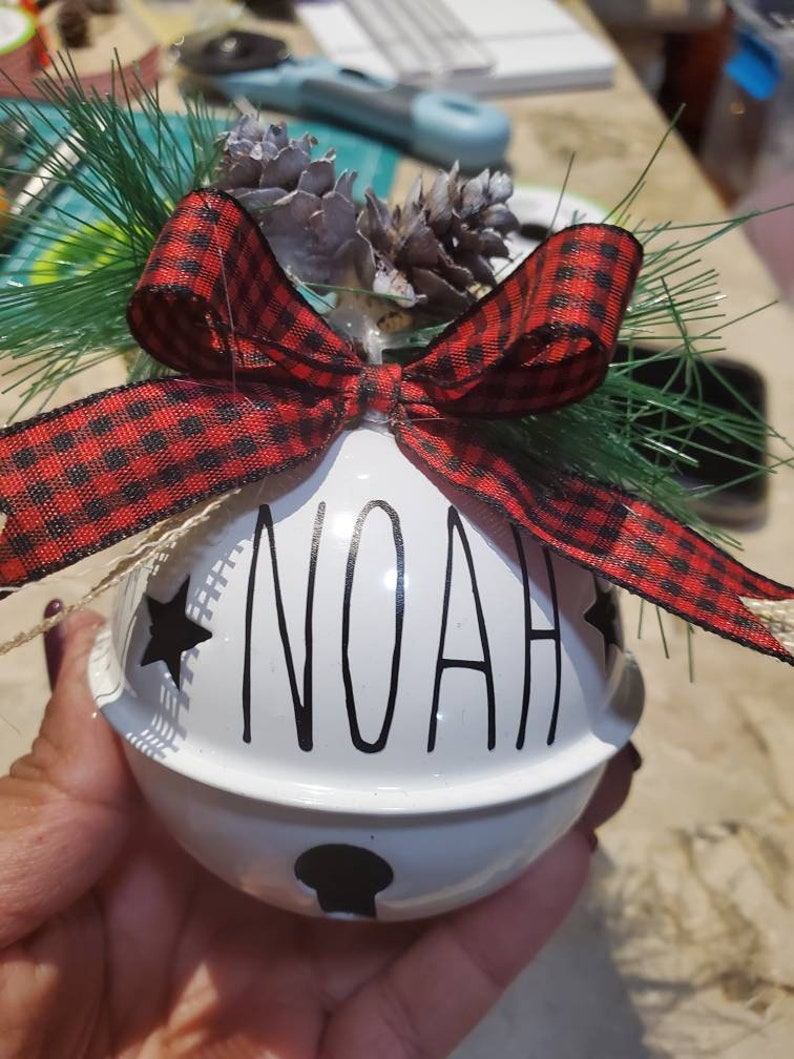 May include: White ceramic jingle bell ornament with the name "NOAH" written in black script. The bell is decorated with a red and black plaid bow and two small pine cones.