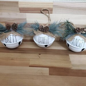 May include: Three white jingle bells with burlap bows and pine sprigs. The bells are personalized with the names "Brett", "Danielle", and "Ainsley".