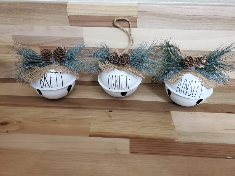 May include: Three white jingle bells with burlap bows and pine sprigs. The bells are personalized with the names "Brett", "Danielle", and "Ainsley".