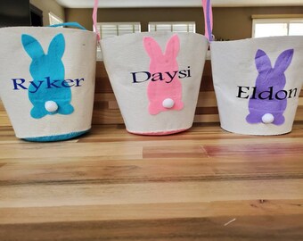 Puede incluir: Tres cestas de Pascua de lona blanca con diseños de conejos. Las cestas están decoradas con conejos de fieltro azul, rosa y morado con pompones blancos para las colas. Los nombres en las cestas son Ryker, Daysi y Eldon.