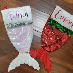 May include: Two personalized mermaid tail Christmas stockings with sequin fabric. The stocking on the left is white with pink sequins and the name "Aubrey" written in purple. The stocking on the right is red with green and red sequins and the name "Emery" written in purple.
