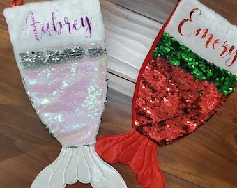 May include: Two personalized mermaid tail Christmas stockings with sequin fabric. The stocking on the left is white with pink sequins and the name "Aubrey" written in purple. The stocking on the right is red with green and red sequins and the name "Emery" written in purple.