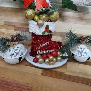 May include: Three white ceramic bells with the names "Danielle", "Ainsley", and "Merry Christmas" written on them. Each bell is decorated with a pine sprig and burlap bow. The bells are sitting on a wooden surface.