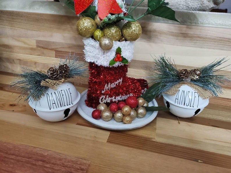 May include: Three white ceramic bells with the names "Danielle", "Ainsley", and "Merry Christmas" written on them. Each bell is decorated with a pine sprig and burlap bow. The bells are sitting on a wooden surface.