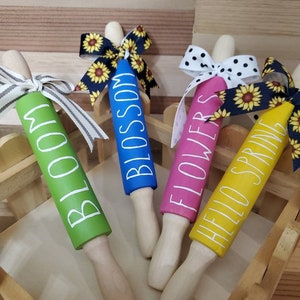 May include: Four wooden rolling pins decorated with spring-themed words and sunflower ribbon. The rolling pins are painted in green, blue, pink, and yellow. The words on the rolling pins are "Bloom", "Blossom", "Flowers", and "Hello Spring".