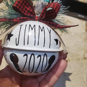 May include: White Christmas ornament shaped like a bell with the name "Jimmy" and the year "2020" written in black. The ornament is decorated with a red and black plaid ribbon, a pine sprig, and a burlap bow.