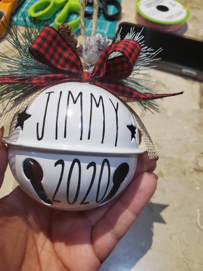 May include: White Christmas ornament shaped like a bell with the name "Jimmy" and the year "2020" written in black. The ornament is decorated with a red and black plaid ribbon, a pine sprig, and a burlap bow.