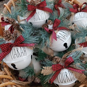 May include: A basket of white ceramic bells with red and black plaid ribbon and pine sprigs. The bells are personalized with the names Ainslie, Alfredo, Mariah, Mari, Brett, and Lourde.