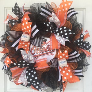 May include: A black, white, and orange wreath with baseball-themed ribbons and a Baltimore Orioles logo. The wreath is decorated with black and white polka dot ribbons, black and white striped ribbons, and orange ribbons with white stripes. The wreath also features a white baseball with the words "Major League Baseball" printed on it.