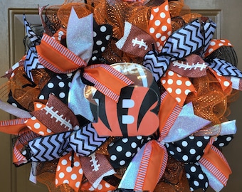 May include: A black, orange, and white wreath with a football helmet in the center. The helmet has the number "13" on it. The wreath is decorated with black and white polka dot ribbon, orange and white striped ribbon, and black and white chevron ribbon.