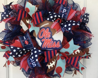 May include: A blue and red Ole Miss wreath with a football theme. The wreath is decorated with red and white polka dot ribbon, blue and red striped ribbon, and a football-shaped sign that says "Ole Miss".