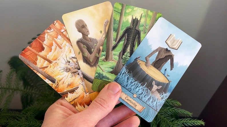 May include: Four tarot cards with illustrations of nature spirits. The cards are held in a hand. The card in the foreground is blue with a tree spirit with a book for a head. The text on the card reads "Curiosity".