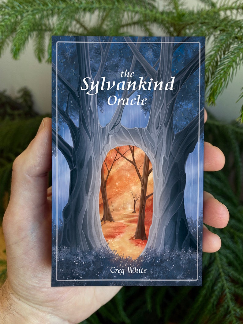 May include: The Sylvankind Oracle deck box features a blue and gray illustration of two trees with a forest scene visible through the opening between the trees. The text "the Sylvankind Oracle" is printed in white at the top of the box. The text "Greg White" is printed in white at the bottom of the box.